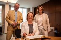 Die Botschafterin sitzt am Konferenztisch im Präsidentinnenzimmer und trägt sich in das Gästebuch ein. Hinter ihr stehen die Präsidentin und der Honorarkonsul. Alle gucken freundlich in die Kamera.